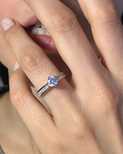 La imagen muestra el anillo "Lumière", con una piedra principal grande en una montura elevada, sostenida por seis garras. Los brazos del anillo están incrustados con piedras de diferentes tamaños, creando un efecto brillante y deslumbrante.